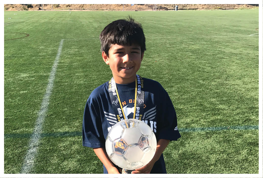 A child wearing a dark sports shirt stands on a grassy field holding a soccer ball and wearing a medal around their neck. Description generated by AI