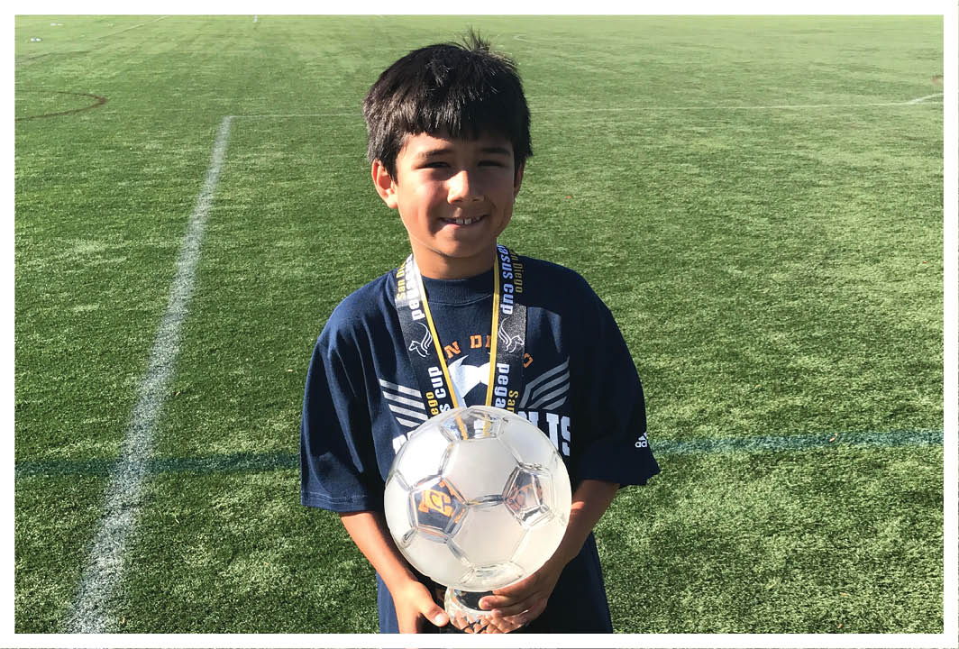 A child wearing a dark sports shirt stands on a grassy field holding a soccer ball and wearing a medal around their neck. Description generated by AI