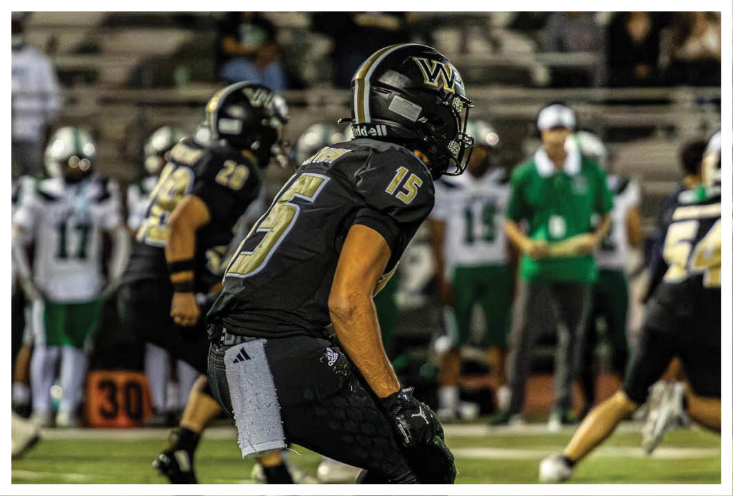 A person wearing a black football uniform with the number 15 is preparing to play on a field with other players and a coach in the background. Description generated by AI