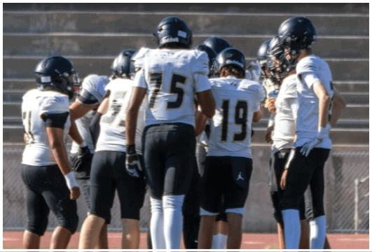 A person wearing a black football uniform with the number 15 is preparing to play on a field with other players and a coach in the background. Description generated by AI