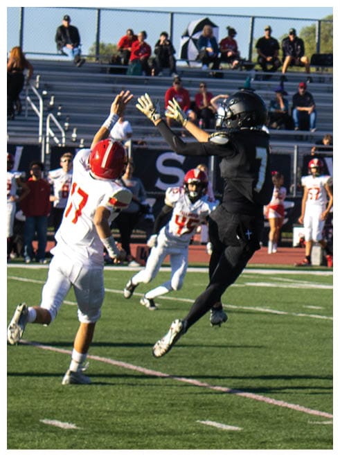 Two people in football uniforms are jumping and reaching for the ball on a field with spectators in the background. Description generated by AI