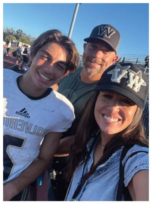 Three smiling people, one wearing a sports jersey and two wearing caps, pose closely together outdoors on a sunny day. Description generated by AI