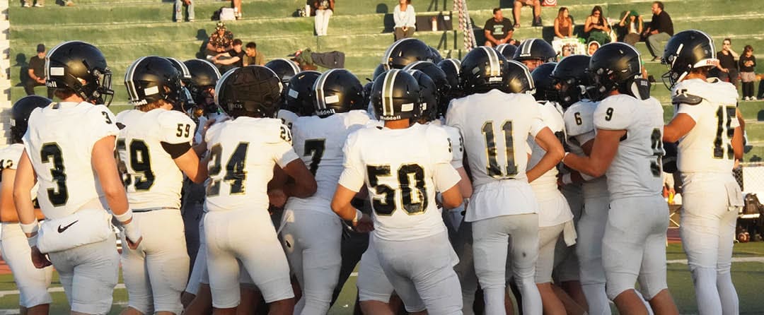 A group of people wearing white football uniforms and black helmets are huddled together on a field with spectators sitting on bleachers in the background. Description generated by AI