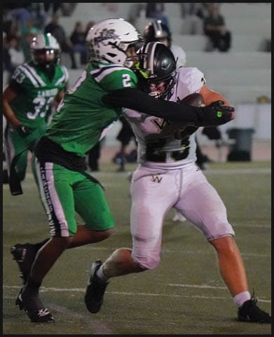 A person in white sports gear is running on a football field while another person in green sports gear is running in the opposite direction with blurred spectators in the background. Description generated by AI