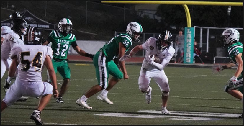 Players in green and white football uniforms are actively engaged in a nighttime game on a field with a scoreboard showing the home team leading. Description generated by AI