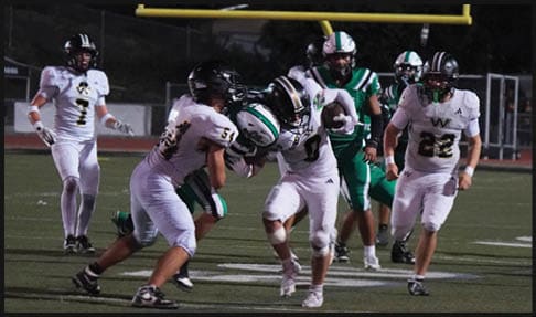 People in football uniforms are actively playing on a field under stadium lights with a scoreboard visible in the background. Description generated by AI