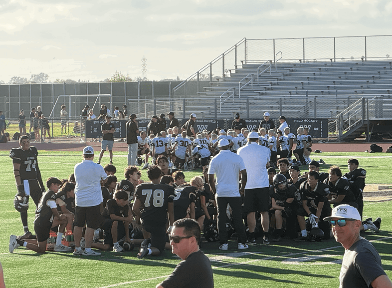 Four people wearing black football uniforms with “VISTA" on the front are standing together in a stadium with a crowd in the background, two of them holding helmets raised above their heads. Description generated by AI