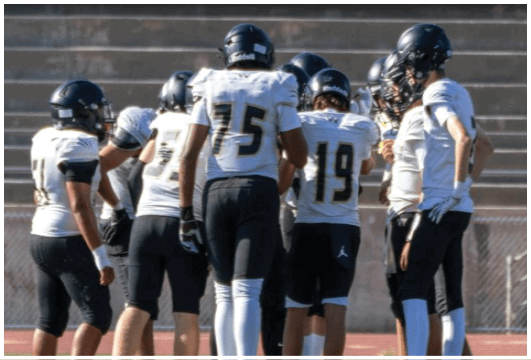 A person wearing a black football uniform with the number 15 is preparing to play on a field with other players and a coach in the background. Description generated by AI
