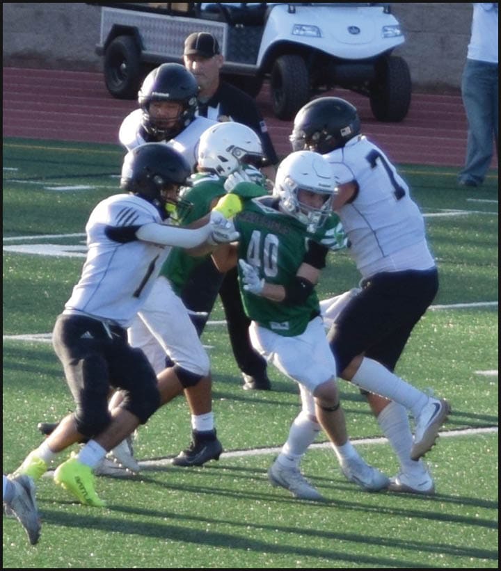 Several people in football uniforms are engaged in a tackle on a green field while a person in casual clothing takes a photo in the background. Description generated by AI