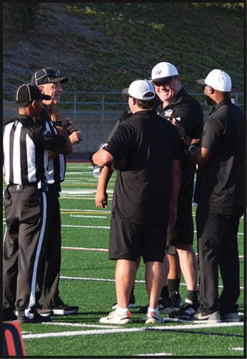 Four people wearing black shirts and white caps are standing on a sports field, engaged in conversation with one person smiling. Description generated by AI