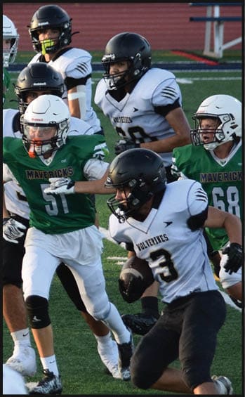Several people wearing football helmets and uniforms are actively engaged in a game on a field, with one person in a green jersey running while others in white jerseys appear to be defending. Description generated by AI