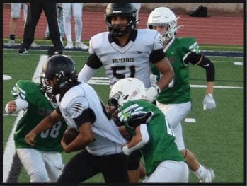 People wearing football helmets and uniforms are actively playing on a football field with a track and a wall in the background. Description generated by AI