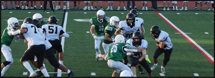 People wearing sports uniforms are actively playing football on a grassy field with a coach or referee in the foreground. Description generated by AI