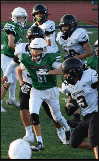 Several people wearing football helmets and uniforms are actively engaged in a game on a field, with one person in a green jersey running while others in white jerseys attempt to block or tackle. Description generated by AI
