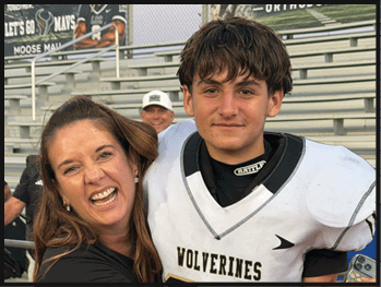 A person in a white football uniform stands next to a smiling person with long hair while a person wearing a white cap smiles in the background on bleachers. Description generated by AI