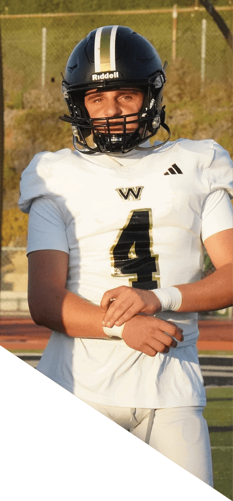 A person wearing a white football uniform with the number four and a black helmet stands on a sports field with a fence and grassy hill in the background. Description generated by AI