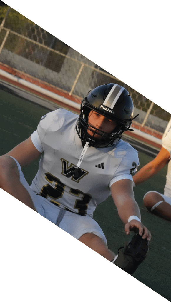 Two people in football uniforms and helmets are sitting on a field with one person stretching their legs. Description generated by AI