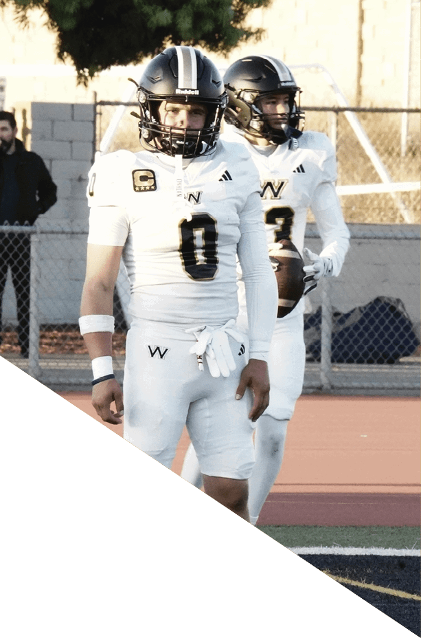 Two people in white football uniforms and helmets stand on a sports field, with one holding a football. Description generated by AI
