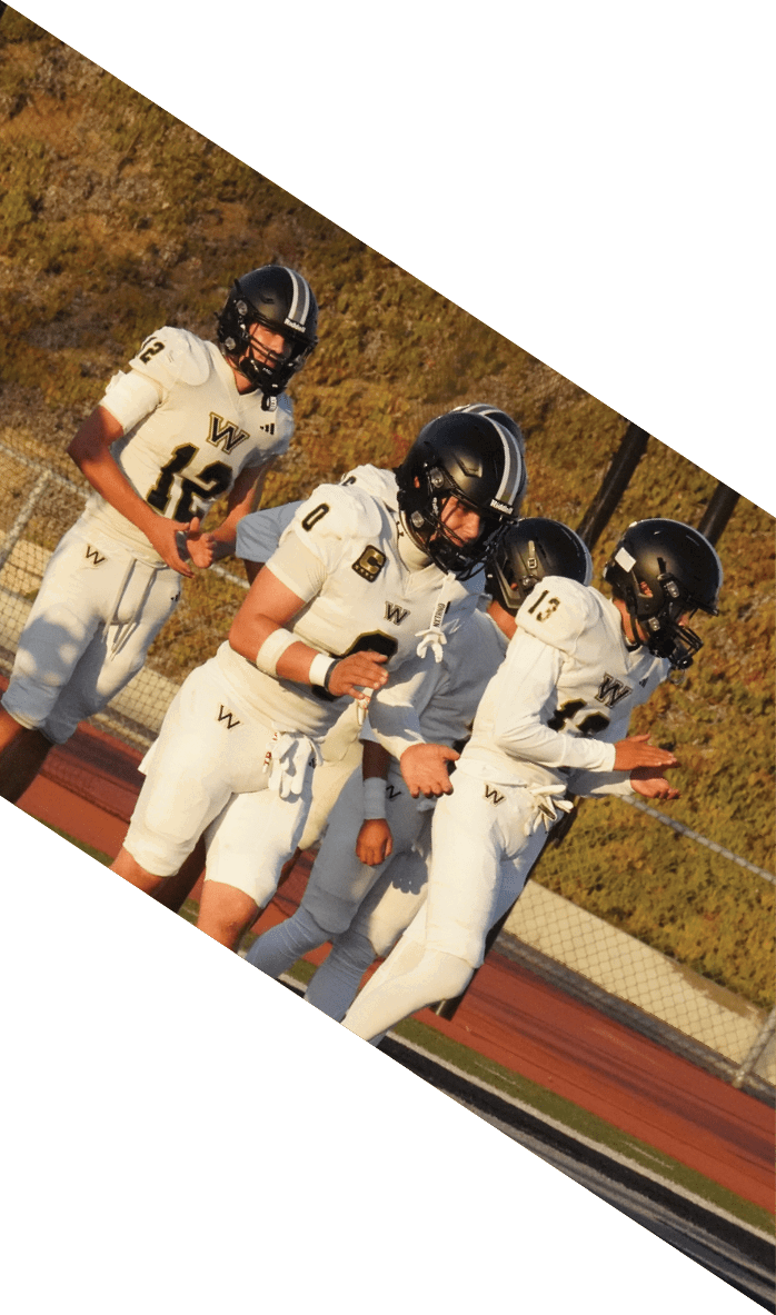 People in white football uniforms with black helmets are standing and preparing on the sidelines of a football field. Description generated by AI