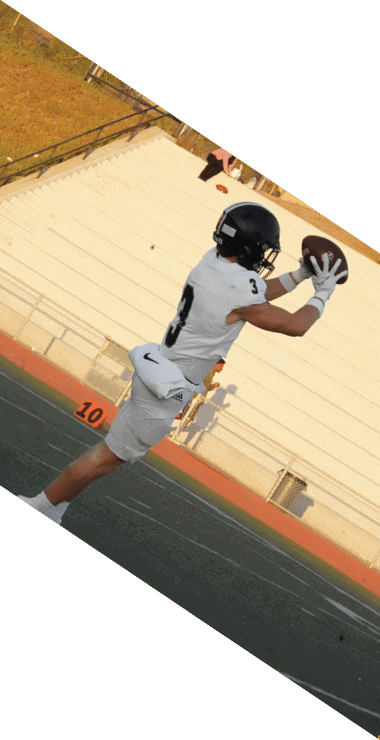 A person in a white football uniform and black helmet is running on a football field near the sideline with empty bleachers and a few people in the background. Description generated by AI