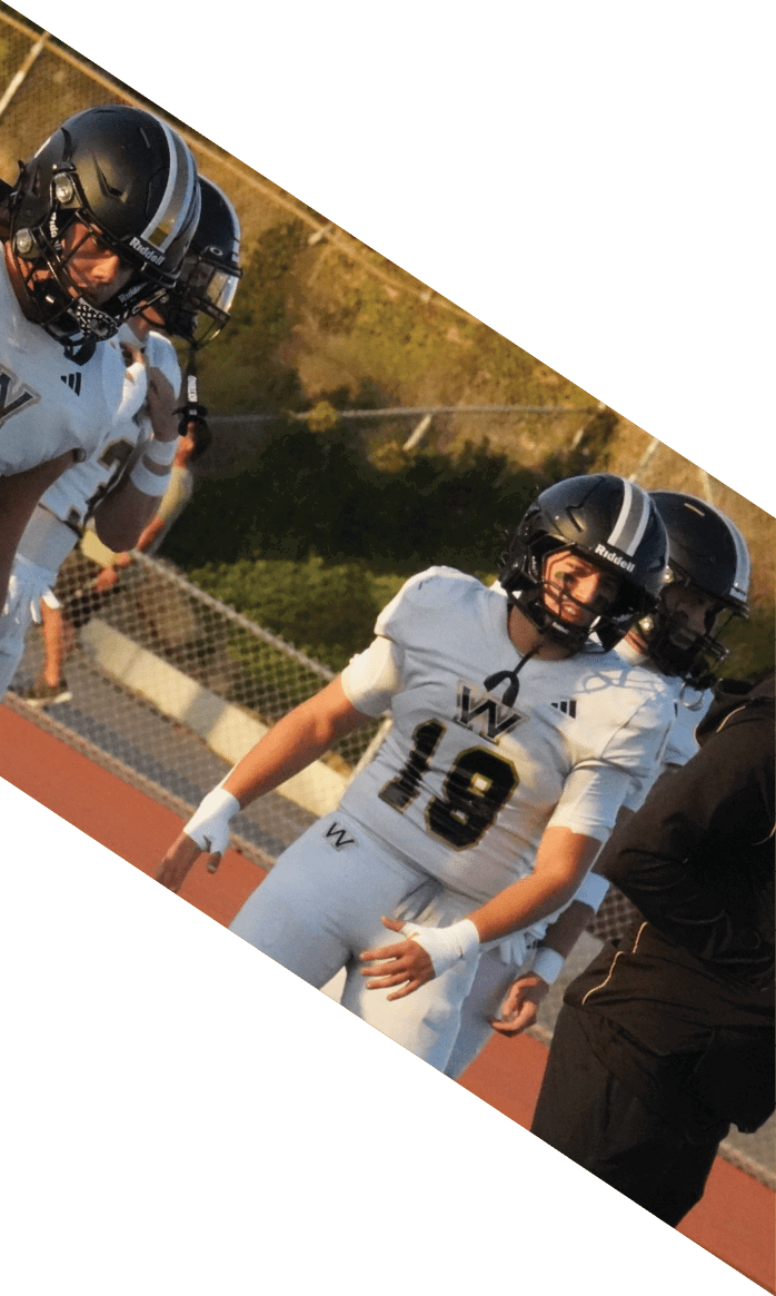 People in white football uniforms and helmets stand on a field during a game or practice. Description generated by AI