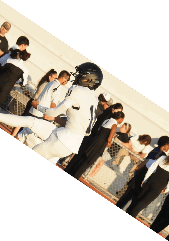A person in white football gear is kicking a ball on a field while several people stand behind a fence watching. Description generated by AI