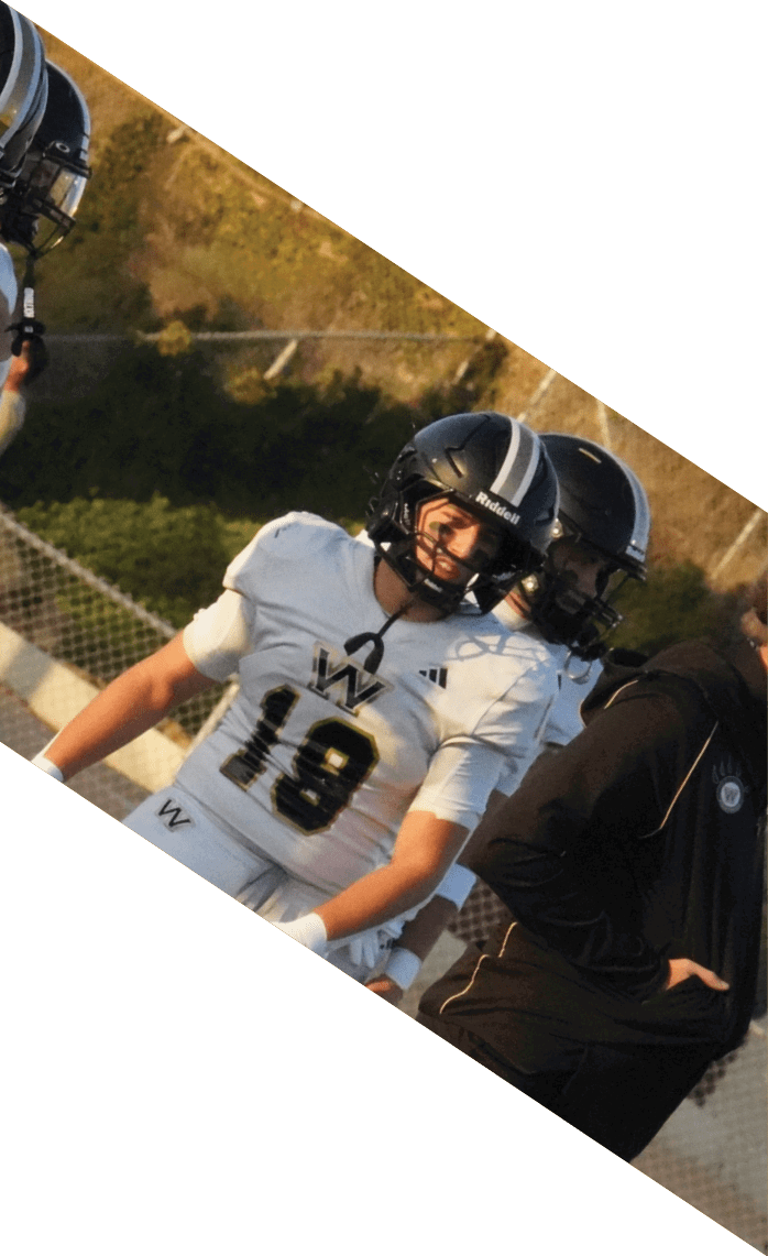 People wearing white football uniforms with black numbers are standing on an outdoor sports field near a chain-link fence. Description generated by AI