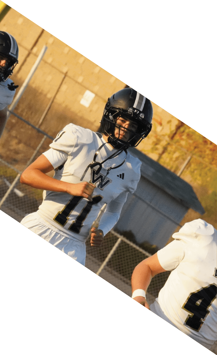 People wearing white football uniforms and black helmets are running on a field near a chain-link fence and a building in the background. Description generated by AI