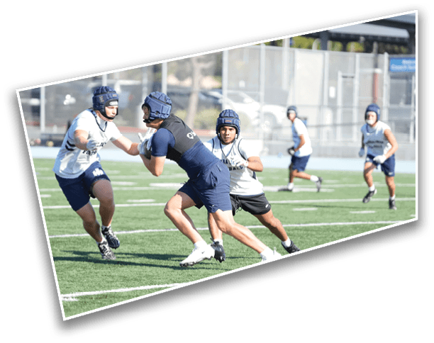 People wearing helmets and athletic gear are actively playing a contact sport on a grassy field with a fenced background. Description generated by AI