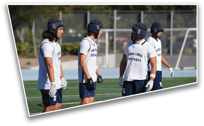 Four people wearing helmets, white shirts with “TURBA LINDA FOOTBALL" text, and dark shorts stand on a sports field. Description generated by AI