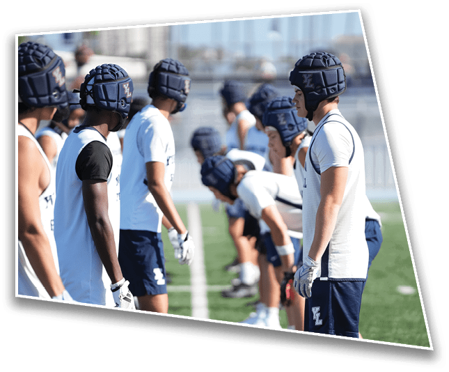 People wearing helmets and athletic clothing stand facing each other in two lines on a sports field, appearing to prepare for a game or practice. Description generated by AI