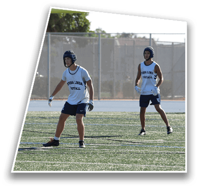 Two people wearing helmets and sports attire stand on a football field under a goalpost, appearing ready to play. Description generated by AI
