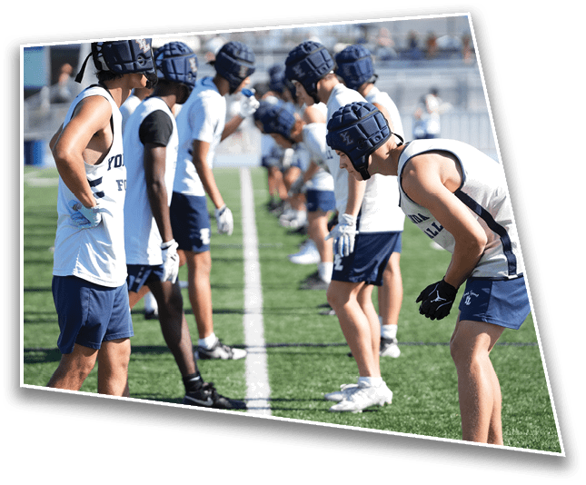 People wearing helmets and athletic clothing stand in two lines facing each other on a sports field, appearing to prepare for a drill or game. Description generated by AI