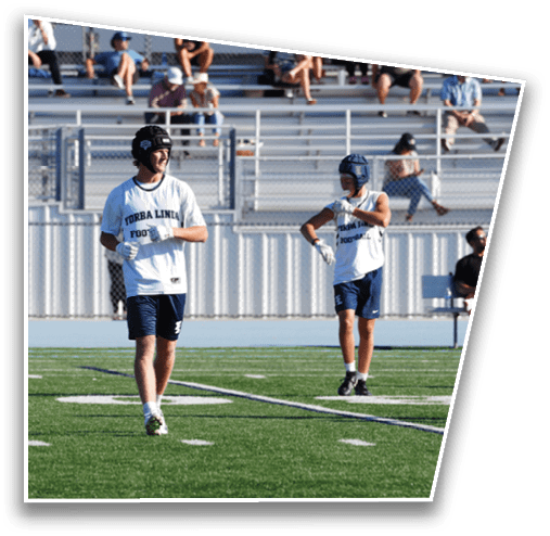A person wearing a sports helmet and a white shirt with text stands on a green football field near the sidelines with empty bleachers in the background. Description generated by AI