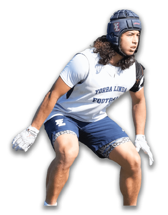A person wearing a football helmet, gloves, a white sleeveless shirt with “Yorba Linda Football" printed on it, and blue shorts is in a crouched athletic stance. Description generated by AI