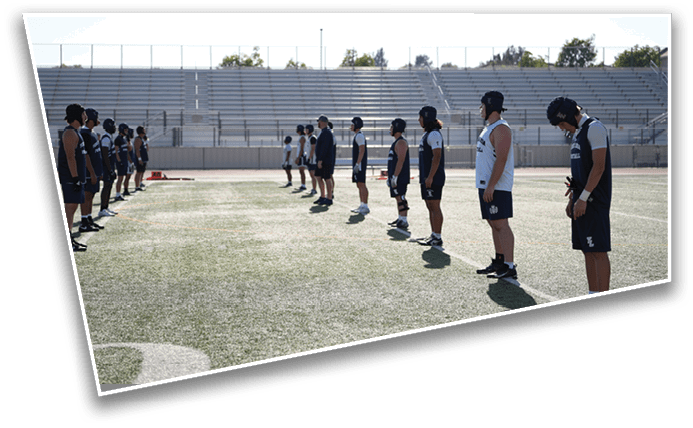 People wearing sports gear and helmets stand in two lines facing each other on a field with empty bleachers in the background. Description generated by AI