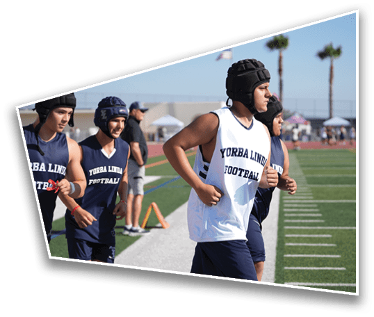 People wearing sports helmets and athletic clothing are running on a track field during a daytime event. Description generated by AI