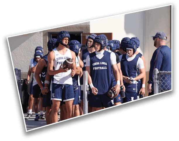 A group of people wearing football helmets and athletic clothing are gathered outdoors near a fence, with one person holding a football and another person standing nearby observing. Description generated by AI