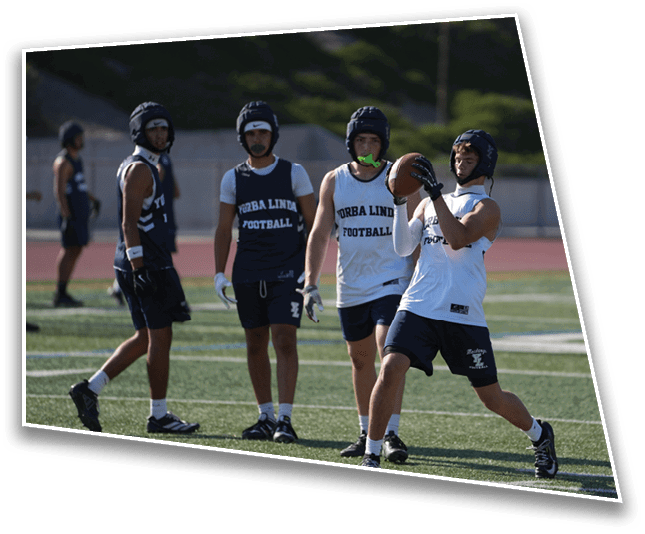 Four people wearing sports helmets and athletic clothing are on a football field, with one person catching a football while the others watch. Description generated by AI
