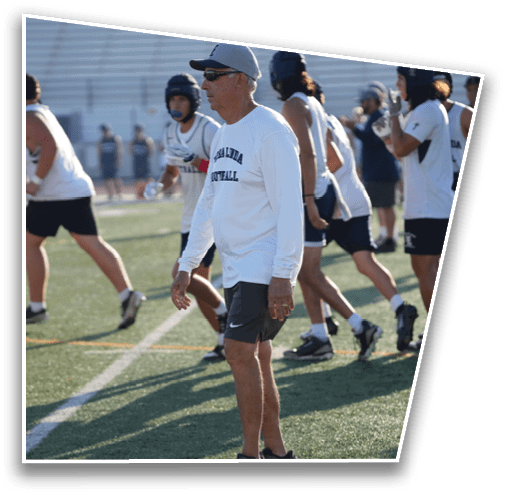 A person wearing a white long-sleeve shirt and black shorts stands on a sports field with other people walking or running in the background. Description generated by AI