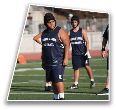 A person wearing a blue sleeveless sports jersey and shorts with a padded helmet stands on a football field while another similarly dressed person stands in the background. Description generated by AI