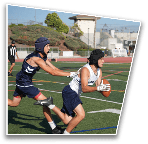 Two people wearing protective headgear are playing flag football on a field, with one person holding the ball while the other reaches to grab a flag. Description generated by AI