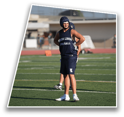 A person wearing a sleeveless shirt and shorts with “TURBA LINDA FOOTBALL" printed on the shirt stands on a football field with hands on hips. Description generated by AI