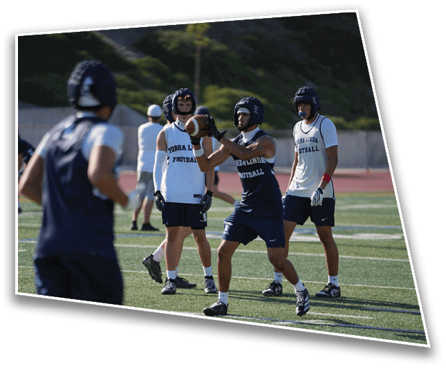 Adults wearing football helmets and practice jerseys are on a sports field with one person preparing to catch a football. Description generated by AI
