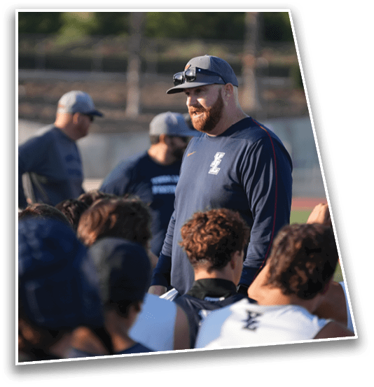 A person wearing a cap and sunglasses on their head is speaking to a group of people outdoors on a sports field. Description generated by AI
