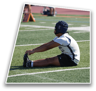 A person wearing athletic gear and a padded helmet is sitting on a sports field stretching forward to touch their toes. Description generated by AI