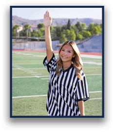 A person with long hair wearing a black and white striped referee shirt is standing on a sports field with one arm raised and smiling. Description generated by AI