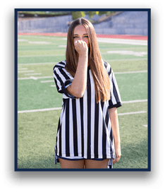 A person with long straight hair wearing a black and white striped shirt stands on a sports field with one hand partially covering their face. Description generated by AI