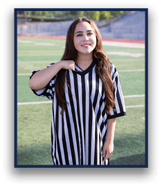 A person with long hair wearing a black and white striped referee shirt stands on a sports field with a slight smile. Description generated by AI