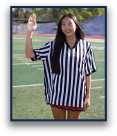 A person with long hair wearing a black and white striped referee shirt is standing on a sports field with one hand raised. Description generated by AI